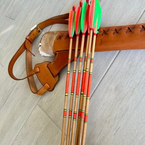 May include: A set of six wooden arrows with red and green fletching and red and green bands. The arrows are in a brown leather quiver with a strap.