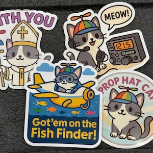 Op de afbeelding: Een verzameling kleurrijke stickers met cartoonkatten. Eén kat is verkleed als priester met de tekst "WITH YOU". Een andere kat zit in een vliegtuig met de tekst "Got'em on the Fish Finder!". Andere stickers bevatten een kat met een radio en de tekst "MEOW!" en "PROP HAT CAT".