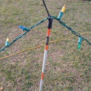May include: A white and orange striped pole with a black cord attached to the top. The cord is connected to a string of Christmas lights with yellow, blue, and green bulbs.