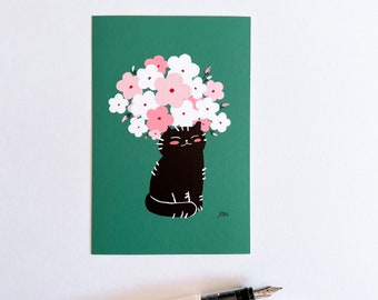 May include: A whimsical art print featuring a black cat with a bouquet of pink and white flowers on its head, set against a teal background. A white fountain pen with a clear cap rests below the print.
