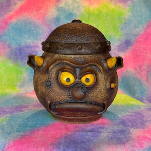 May include: A brown ceramic jar with a grumpy face, yellow eyes, and a textured surface. The jar has a lid with a metal band and rivets.