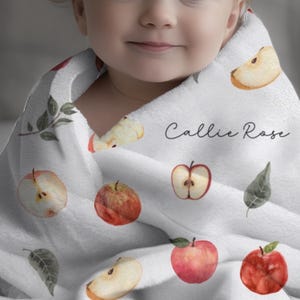 May include: A white baby blanket with a watercolor apple and leaf pattern. The blanket has the name "Callie Rose" written on it.