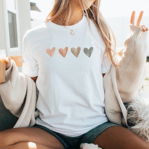 May include: White t-shirt with four watercolour hearts in shades of pink, orange, brown, and green.