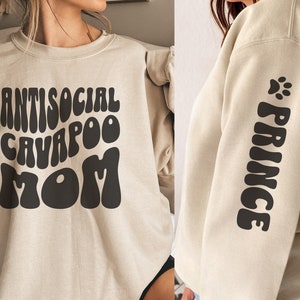 Könnte beinhalten: Ein beiger Pullover mit dem Text "ANTISOCIAL CAVAPOO MOM" in schwarzen Retro-Lettern auf der Vorderseite. Der passende Ärmel hat den Text "PRINCE" in schwarzen Retro-Lettern mit einem Pfotenabdruck darüber.
