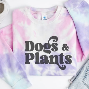 May include: A tie-dye sweatshirt with a pink, purple, and blue color scheme. The sweatshirt has the text "Dogs & Plants" in black retro-style lettering.