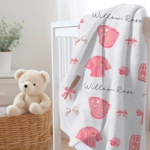 May include: White baby blanket with a pink baseball theme. The blanket features pink baseballs, bats, gloves, and jerseys. The words "Willow Rose" are printed on the blanket.