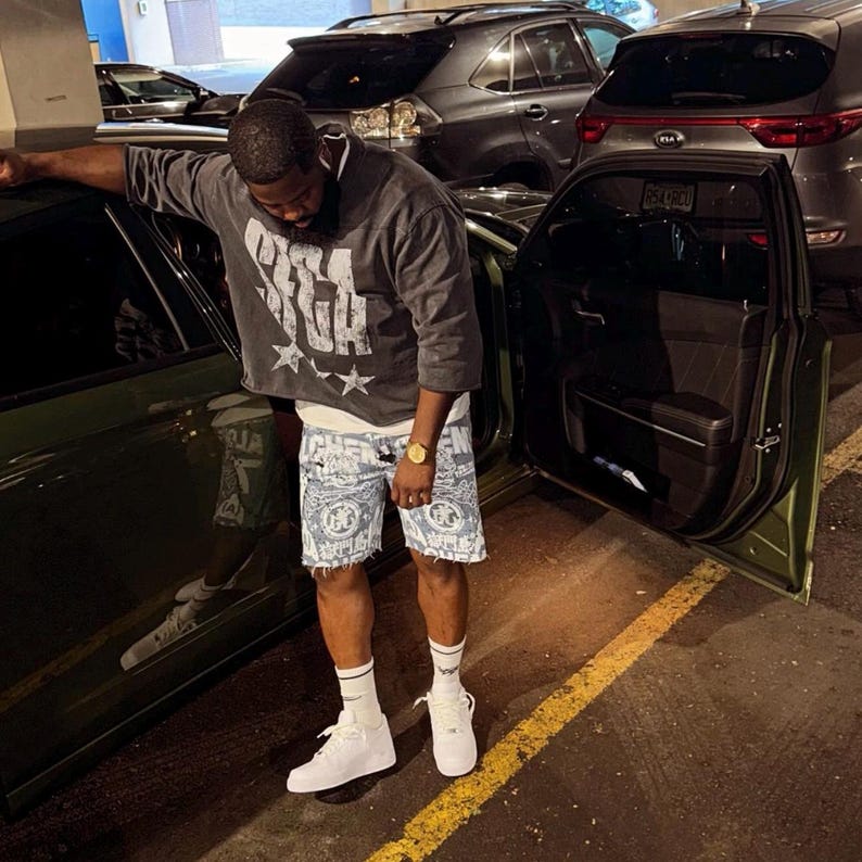 May include: A person wearing a gray cropped sweatshirt with white text and two stars, paired with blue and white patterned shorts. The person is wearing white sneakers and a gold watch. The car door is open.