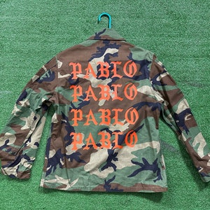 Könnte beinhalten: Eine Jacke in Tarnfarben mit Grün und Braun, auf der Rückseite steht viermal das Wort "PABLO" in orangefarbener Schrift.