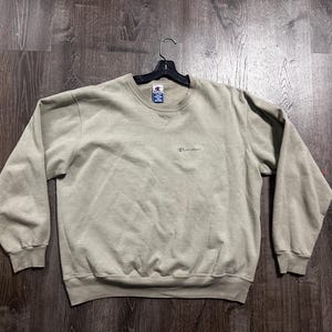 May include: A light green Champion sweatshirt with a crew neck and long sleeves. The Champion logo is embroidered in a small font on the chest. The sweatshirt is hanging on a black hanger.