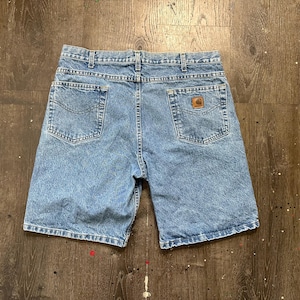May include: Light blue denim shorts featuring a classic five-pocket design, a button and zipper fly closure, and belt loops. A small rectangular brown leather-like patch with the Carhartt logo is stitched onto the back right pocket.
