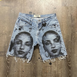 May include: Light blue denim shorts with a distressed hem. The shorts feature a black and white graphic of a woman's face on each leg, with black paint splatter details. The shorts are a Wrangler brand.