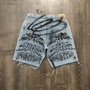 May include: Light blue denim shorts with distressed edges. The shorts feature black spray-painted angel wings and the word "PHNTM" in an oval design on the back. A black halo is also spray-painted on the back.