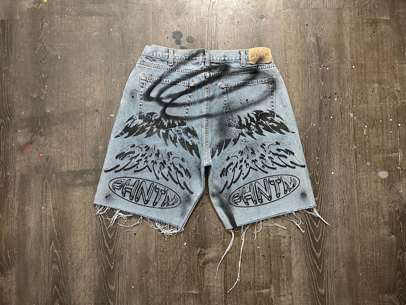 May include: Light blue denim shorts with distressed edges. The shorts feature black spray-painted angel wings and the word "PHNTM" in an oval design on the back. A black halo is also spray-painted on the back.