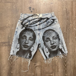 May include: Light blue denim shorts with a distressed hem. The shorts feature a black and white graphic of a woman's face with hoop earrings and a honeycomb pattern on the face. The shorts are on a wooden surface.