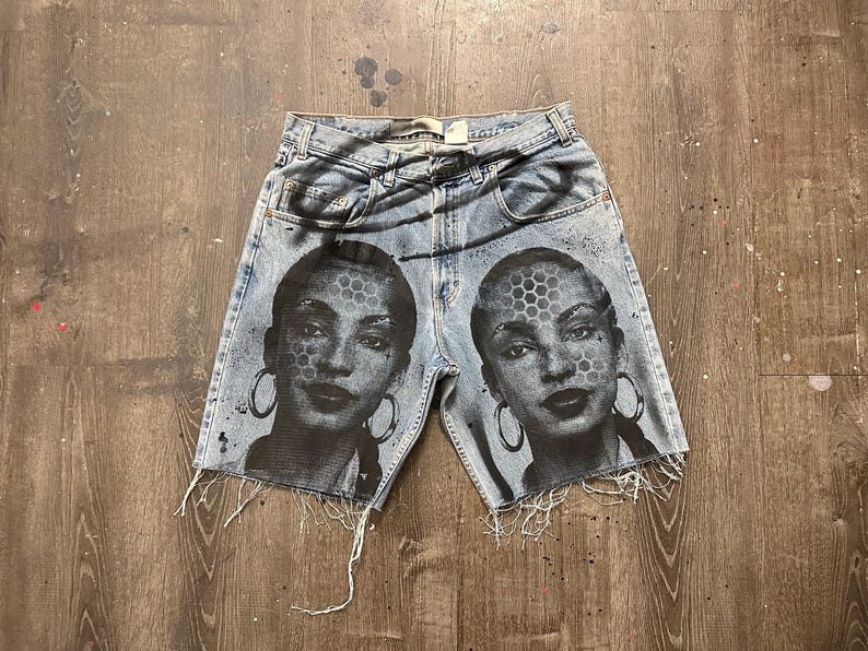May include: Light blue denim shorts with a distressed hem. The shorts feature a black and white graphic of a woman's face with hoop earrings and a honeycomb pattern on the face. The shorts are on a wooden surface.