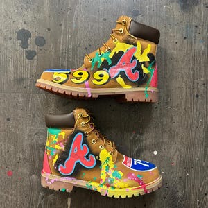 May include: A pair of tan leather ankle boots featuring custom graffiti-style paint splatters in yellow, pink, and teal. The boots display a red letter A logo, the number 599, and a blue Interstate 75 road sign graphic with yellow laces.