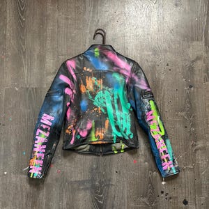 Puede incluir: Chaqueta de cuero negra con un vibrante diseño de pintura en aerosol en rosa, azul, verde y naranja. Las mangas presentan la palabra "MEZANINE" en rosa. La chaqueta está colgada en una percha negra sobre un fondo de madera.