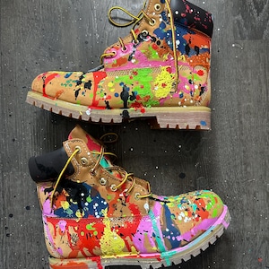 May include: A pair of tan leather ankle boots featuring a vibrant multi-coloured paint splatter design. The boots have black padded collars, yellow and brown laces, gold-tone metal eyelets, and thick, light-coloured lugged rubber outsoles.