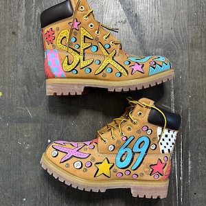 Puede incluir: Un par de botas de trabajo color canela con arte de graffiti colorido. Las botas presentan la palabra "SEX" y el número "69" en amarillo y azul, junto con estrellas y otros diseños. Las botas tienen cuellos negros y cordones amarillos.