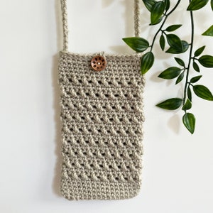 May include: A beige crocheted crossbody bag with a wooden button closure. The bag is hanging from a white wall with a green plant in the background.