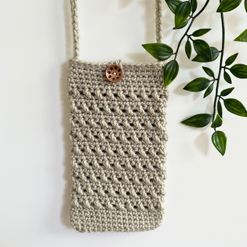 May include: A beige crocheted crossbody bag with a wooden button closure. The bag is hanging from a white wall with a green plant in the background.