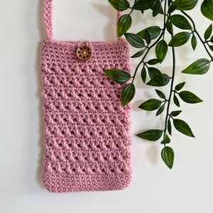 May include: A pink crocheted crossbody bag with a wooden button closure. The bag is hanging from a white wall with green leafy vines in the background.