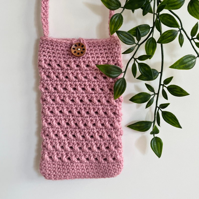 May include: A pink crocheted crossbody bag with a wooden button closure. The bag is hanging from a white wall with green leafy vines in the background.