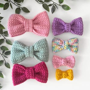 Op de afbeelding: Set van zeven gehaakte strikken in verschillende kleuren, waaronder roze, paars, groen, geel en regenboog. De strikken zijn gerangschikt op grootte, met de grootste strik bovenaan en de kleinste strik onderaan.