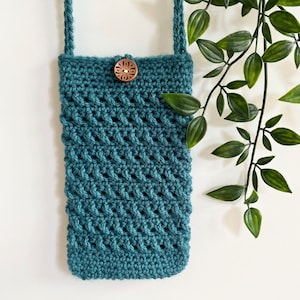 May include: A teal crocheted crossbody bag with a wooden button closure. The bag is hanging from a white wall with green leafy plants in the background.