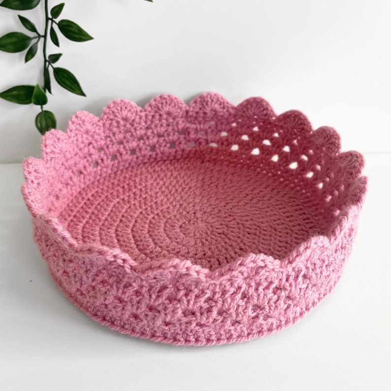 May include: A pink crocheted basket with a scalloped edge. The basket is made with a delicate stitch pattern and has a round shape.