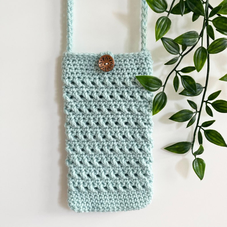 May include: A light blue crocheted phone pouch with a wooden button closure. The pouch is hanging from a light blue crocheted strap.