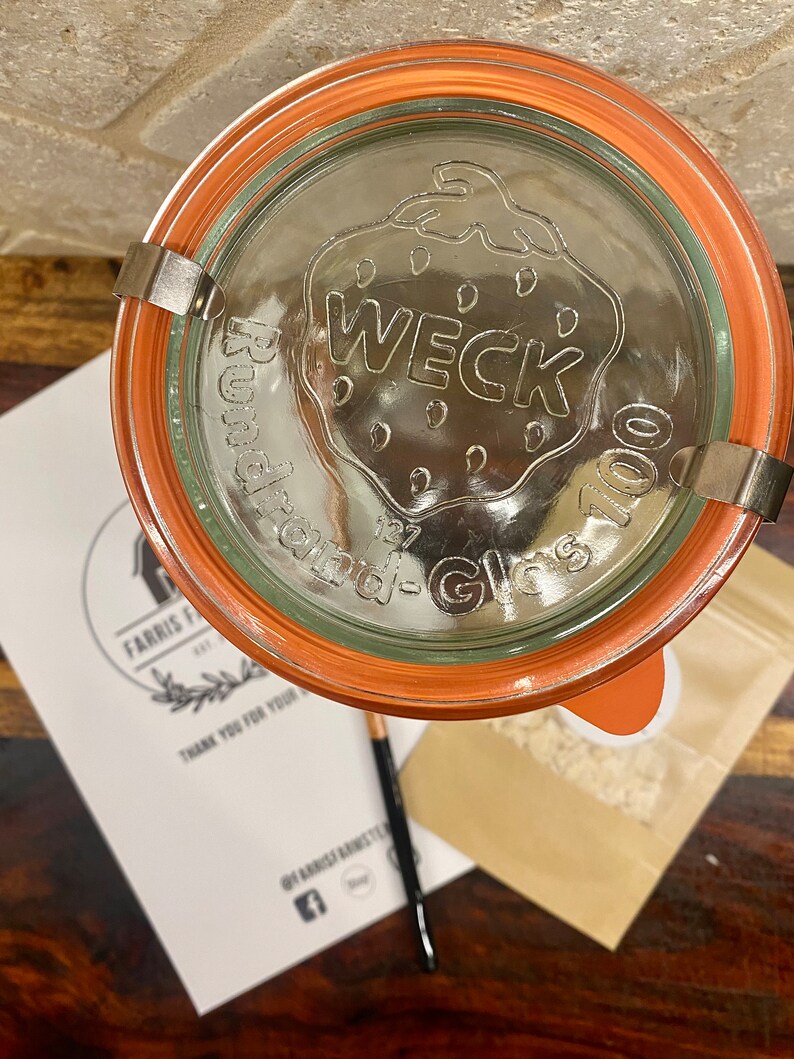 May include: A close-up of a clear glass jar with an orange lid. The lid has a WECK logo and the words "Rundrand-Glas 100".