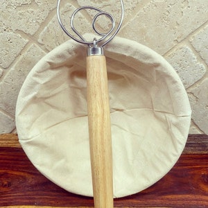 Puede incluir: Un batidor de masa de acero inoxidable con mango de madera descansa sobre una cesta de fermentaci&oacute;n redonda de lino blanquecino. El batidor presenta un dise&ntilde;o en bucle. La cesta est&aacute; vac&iacute;a. Los art&iacute;culos est&aacute;n sobre una superficie de madera.
