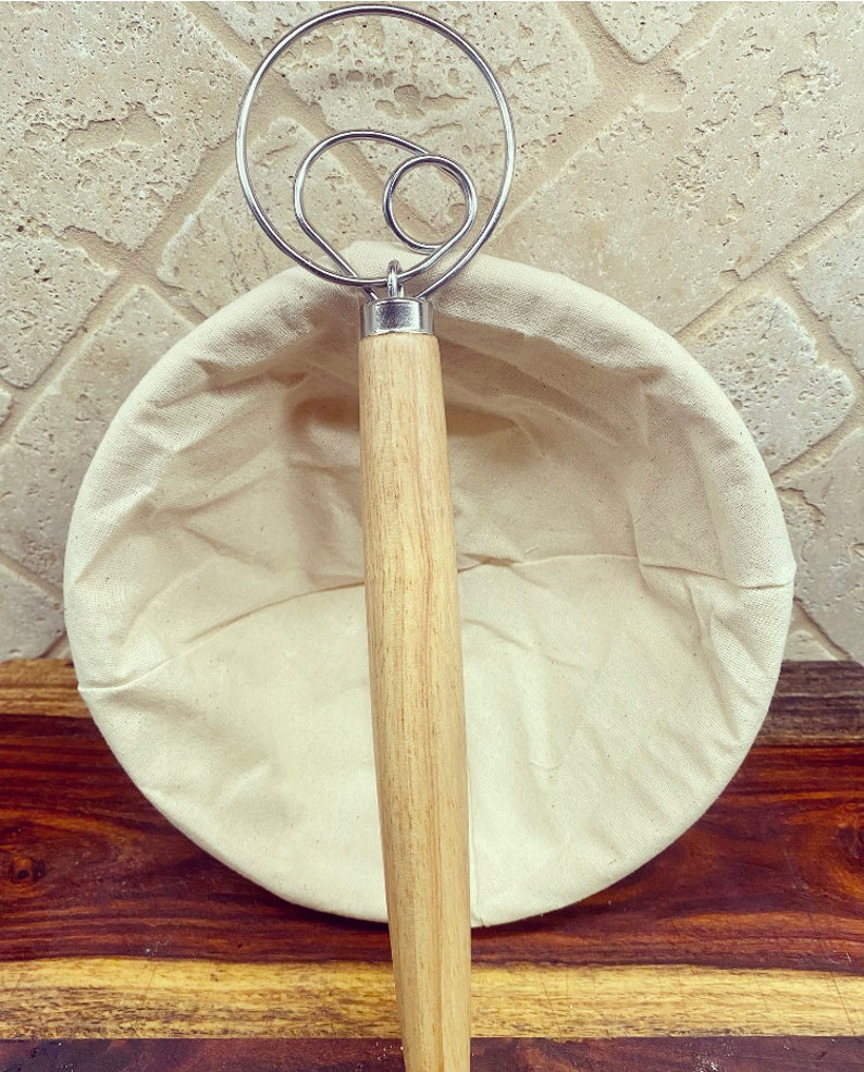 Puede incluir: Un batidor de masa de acero inoxidable con mango de madera descansa sobre una cesta de fermentaci&oacute;n redonda de lino blanquecino. El batidor presenta un dise&ntilde;o en bucle. La cesta est&aacute; vac&iacute;a. Los art&iacute;culos est&aacute;n sobre una superficie de madera.