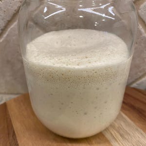 Puede incluir: Un tarro de vidrio lleno de masa madre de pan de masa madre blanca y burbujeante. La masa madre est&aacute; en proceso de fermentaci&oacute;n y tiene una textura espumosa.