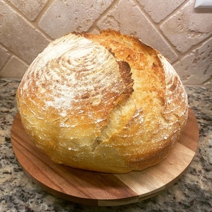 Puede incluir: Una hogaza redonda de pan reci&eacute;n horneado, de color marr&oacute;n dorado con una corteza crujiente, se encuentra sobre una tabla de madera. La superficie del pan est&aacute; espolvoreada con harina, y la textura sugiere un estilo r&uacute;stico y artesanal. El pan es una pieza central, listo para servir.