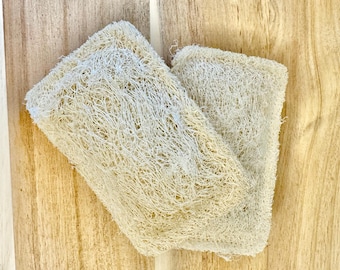 May include: Two natural loofah sponges, beige in color, are stacked on top of each other on a wooden surface.