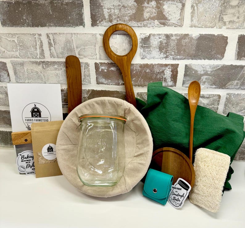 Puede incluir: Un kit de masa madre con un frasco de vidrio, una cesta de fermentaci&oacute;n de lino, una cuchara de madera, una rasqueta de masa de madera, una bolsa de tela verde, una tabla de cortar de madera, una peque&ntilde;a bolsa turquesa, una bolsa de papel marr&oacute;n con el texto "Baking Dish" y una esponja blanca.