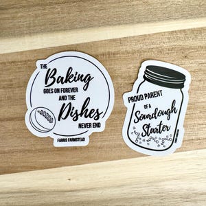 Puede incluir: Dos pegatinas blancas con texto negro y gr&aacute;ficos. La primera pegatina dice "The Baking Goes On Forever And The Dishes Never End" con un peque&ntilde;o gr&aacute;fico de una hogaza de pan. La segunda pegatina dice "Proud Parent Of A Sourdough Starter" con un gr&aacute;fico de un frasco con burbujas.