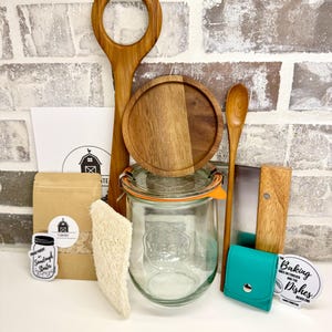 Puede incluir: Un conjunto de art&iacute;culos de cocina que incluye una cuchara de madera, una tabla de cortar de madera, un frasco de vidrio con tapa, una esp&aacute;tula de madera, una bolsa verde, una esponja natural y una pegatina que dice "Los platos para hornear nunca terminan".