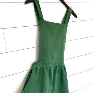 Puede incluir: Un delantal de lino verde con tirantes ajustables y falda fruncida. El delantal tiene un solo bolsillo en la parte delantera.
