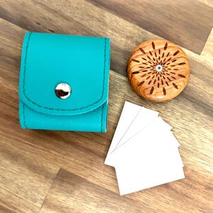 May include: A teal leather case with a silver button closure, a wooden disc with a carved sunburst design, and a stack of white cards.