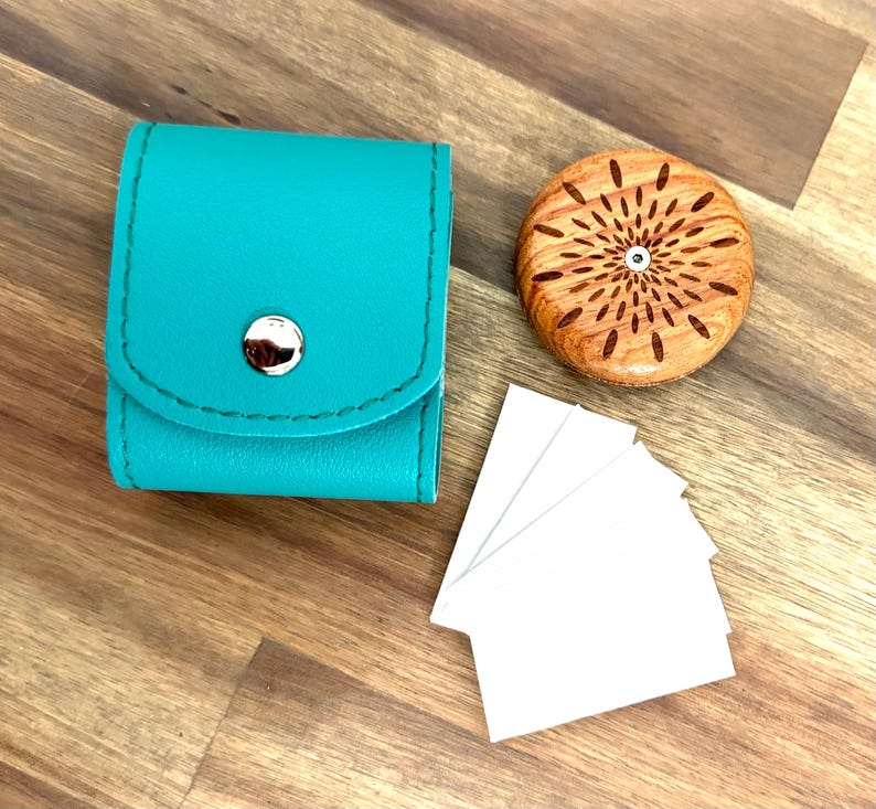 May include: A teal leather case with a silver button closure, a wooden disc with a carved sunburst design, and a stack of white cards.