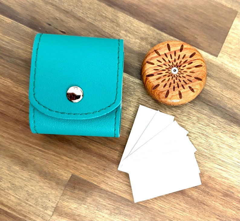 Puede incluir: Un estuche cuadrado de polipiel turquesa con cierre de bot&oacute;n a presi&oacute;n plateado, un objeto redondo de madera con dise&ntilde;o de rayos de sol y una peque&ntilde;a pila de tarjetas rectangulares blancas est&aacute;n sobre una superficie de madera.