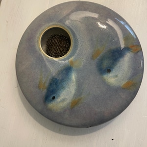 May include: A round, ceramic flower frog with a light blue glaze and two painted blue fish with yellow accents. The frog has a central opening with a metal floral pin holder.