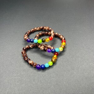 Op de afbeelding: Twee armbanden met kralen in regenboogkleuren. De armbanden zijn gemaakt van kleine, ronde kralen in bruin, rood, oranje, geel, groen, blauw en paars. De armbanden zijn in een achtvorm gerangschikt tegen een donkere achtergrond.