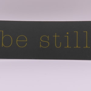 May include: A black rectangular sticker with the words "be still" in gold lettering.