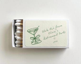 May include: A white matchbox containing matches with white tips. The label features a green line drawing of a martini glass with a bow and the text: Stole this from Olivia's Retirement Party, a small heart symbol, and the year 2026 printed in green.