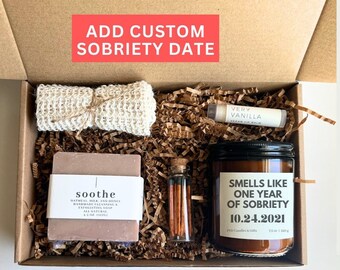 May include: A personalized spa gift box with a candle, soap, matches, and lip balm. The candle reads "Smells Like One Year of Sobriety 10.24.2021." The box includes a cotton soap pouch and a bar of "soothe" soap. The text "Add Custom Sobriety Date" is at the top.
