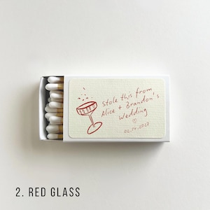 May include: A matchbox with a cream-colored label featuring a red illustration of a champagne glass and the text "Stole this from Alice + Brandon's Wedding 02.14.2027". The match heads are white with wooden stems.